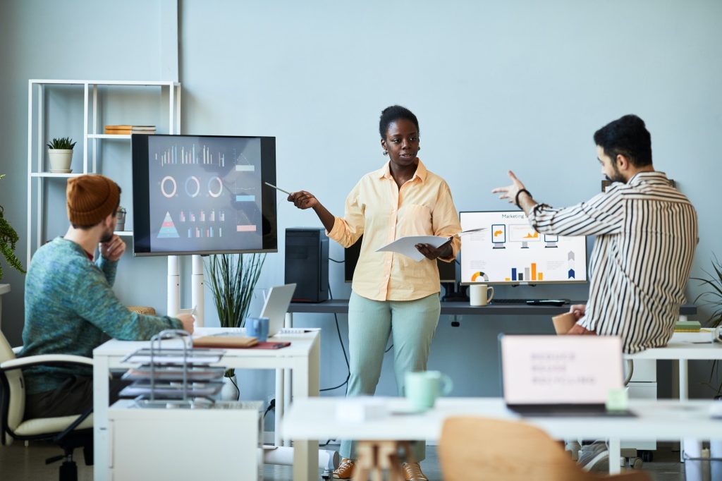 Young confident female business coach pointing at screen with graphic data